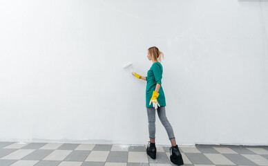 A young girl is engaged in repairs and paints a white wall with a roller in a new apartment. Renovation of the interior and a new apartment. Housewarming and a desirable mortgage.