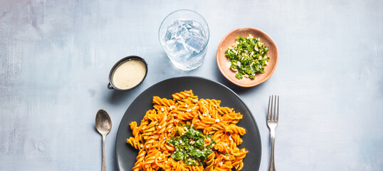 Curly pasta macaroni with tomato and gremolata sauce in a round dark plate on a table with cutlery and a glass of water. Ready to eat.