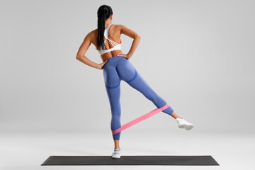 Fitness woman doing exercise for glutes with resistance band on gray background. Athletic girl...