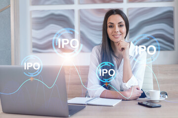 Attractive businesswoman in white shirt at workplace working with laptop to optimize IPO strategy at corporate fund. Financial chart hologram over office background