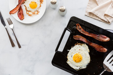 Top down view of bacon and eggs on a griddle pan and on a plate for breakfast.