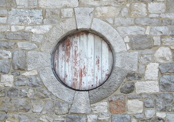 Petite fenêtre ronde nichée sous le toit d'une habitation traditionnelle en pierre, avec volet en bois fermé