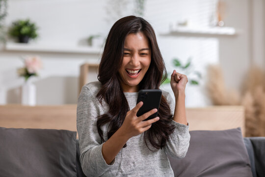 Millennial asian young woman looking mobile phone laughing with good news or discount voucher for shopping online at home.Happy and cheerful woman looking on cellphone app read message feel excited