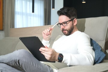 Portrait of a serious handsome bearded guy, young man in glasses is using his tablet laptop computer, relaxing at home on sofa couch, looking at screen, reading electronic book with electronic pen