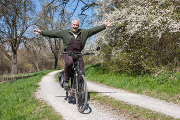 Funny senior man rides his bike hands-free on a dirt road, past flowering sloe bushes and laughs...