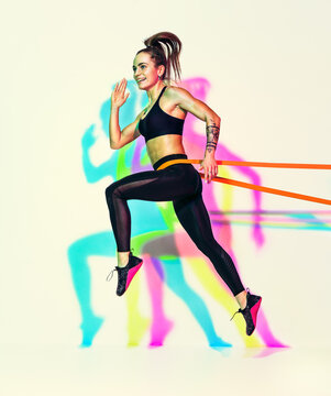 Sporty woman doing jumping exercise with resistance band. Photo of muscular woman in black sportswear on white background with effect of rgb colors shadows.