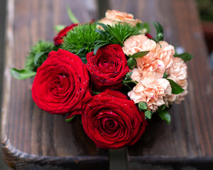 A small bouquet of red roses, delicate pink carnations and green leaves, on a wooden brown background view from above