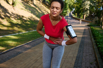 Fototapeta premium Female runner feeling exhaustion and side cramps, stomach-ache during running in the city park. Healthy active lifestyle concept, sport injury