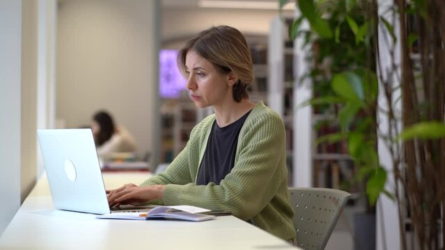 Concentrated female university professor sitting in front of laptop in empty library writing down notes while preparing for lecture or checking course schedule, selective focus. Academic career