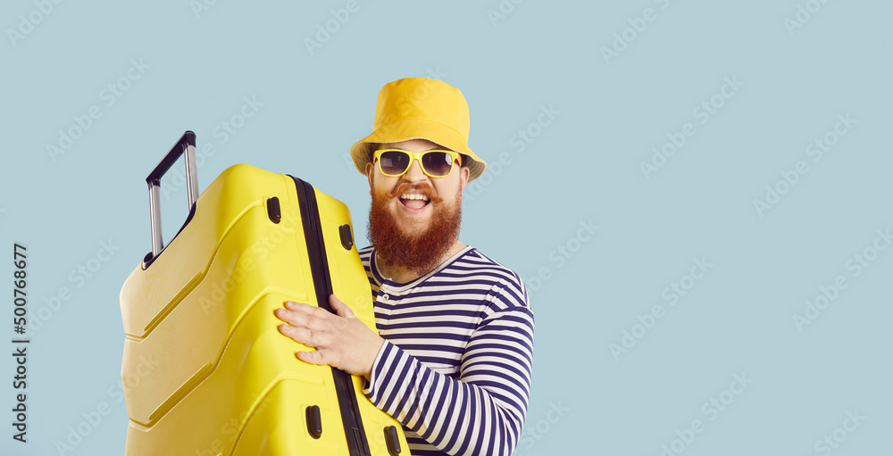 Wall mural Funny fat man going on vacation. Cheerful traveler leaving for summer holiday. Portrait of happy excited bearded guy in striped swimsuit, bucket hat, and sunglasses holding yellow suitcase and smiling