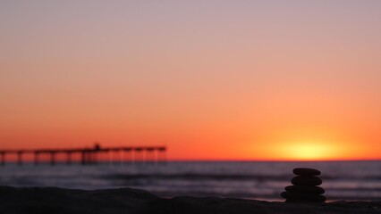 Fototapeta premium Stack of pebble stones, sandy ocean beach, sunset sky. Rock balancing in sun light, sea water waves. Stones staking in pyramid pile. Zen meditation and harmony in balance. Calm tranquil atmosphere.
