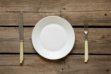 One white plate and a fork with a knife lie on an old wooden table in the kitchen at home, restaurant business