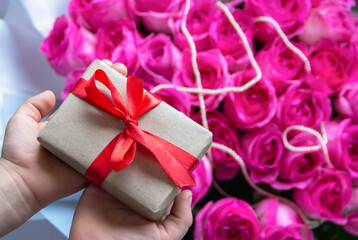 A gift with a red satin ribbon in children's hands against a background of a bouquet of pink roses