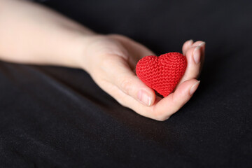 Red knitted heart in palm of female hand on a bed with black bedding. Concept of love, health care,...