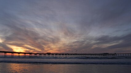 Naklejka premium Silhouette of people walking, pier on piles in sea water. Ocean waves, dramatic sky at sunset. California coast aesthetic, beach or shore vibe at sundown. Summer seascape in San Diego near Los Angeles