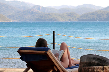 Woman in bikini reading book sitting in deck chair on a sea beach. Summer relax on mountain coast background