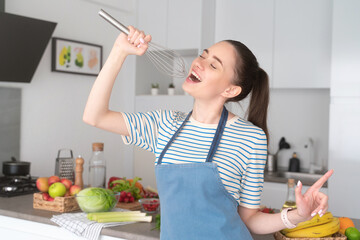 Young fun woman singing a song while cooking food at kitchen
