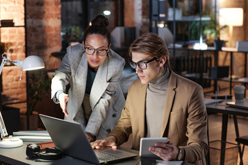 Confident businesswoman in jacket pointing at laptop screen while explaining report to young...