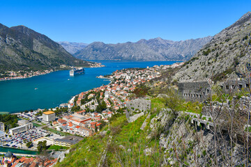 Kotor, Montenegro, Europe. Bay of Kotor on Adriatic Sea. Rock, historical walls, roofs of the buildings in the old town. Cruise in the bay, mountains in the background. Clear blue sky, sunny day