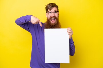 Redhead man with beard isolated on yellow background holding an empty placard with happy expression and pointing it