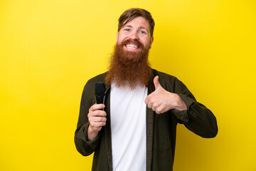 Redhead man with beard picking up a microphone isolated on yellow background with thumbs up because...