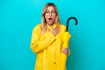 Young Uruguayan woman with rainproof coat and umbrella isolated on blue background surprised and shocked while looking right