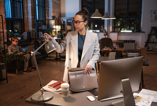 Serious young mixed race businesswoman in glasses standing with bag at table and turning off light in office