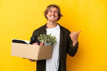 blonde man making a move while picking up a box full of things isolated on yellow background pointing to the side to present a product