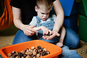 Sensory Play for Kids with Special Needs. Help and Activities for kids with disabilities, Cerebral Palsy. Boy with Cerebral Palsy playing with natural materials cones and nuts - obrazy, fototapety, plakaty