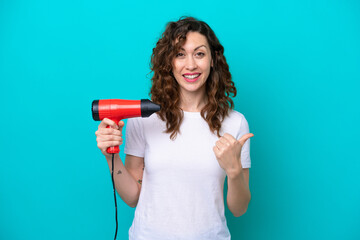 Young caucasian woman holding a hairdryer isolated on blue background pointing to the side to present a product