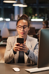 Overworked young woman in glasses sitting at desk and texting sms on smartphone in dark office