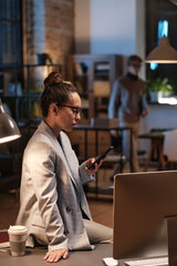 Stylish young mixed race businesswoman in eyeglasses sitting on desk with computer and using smartphone in office at night