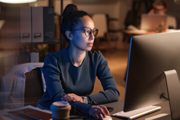 Serious young business lady in glasses sitting at table and using computer while preparing presentation at night