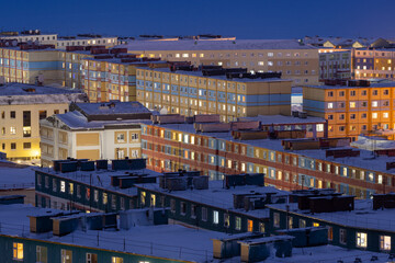 Fototapeta premium Night view from above on a snow-covered northern city in the Arctic. Snow on the roofs of colorful buildings. Cold winter weather. Harsh polar climate. Anadyr, Chukotka, Siberia, Far North of Russia.
