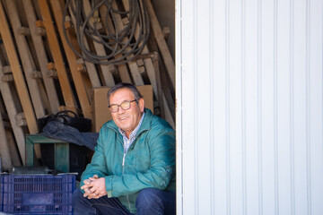 Farmer rests sitting on a box in his farmland's storehouse and smiles looking at the camera