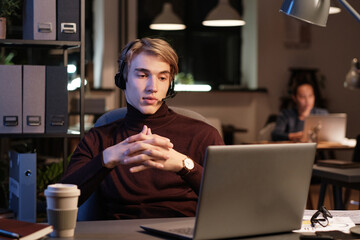 Serious young customer service operator in wireless headset clasing hands while reading online message on laptop while working late