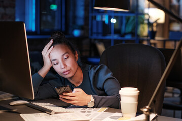 Exhausted young biracial woman sitting at table and leaning head on hand while checking phone message in office at night