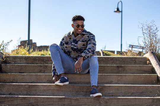 portrait of a smiling young latin man with sunglasses sitting on the street in summertime