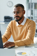 Content young mixed race statistics expert with beard sitting at desk with documents and using laptop in office