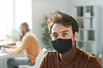 Fototapeta na wymiar Portrait of young mixed race manager of company with brown hair wearing brown turtleneck and black mask in office
