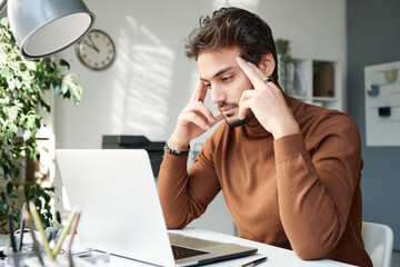 Serious young middle eastern man in brown turtleneck focused on thoughts looking at laptop screen and massaging temples