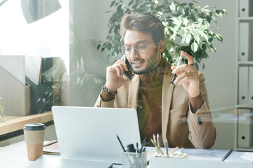 Content confident young mixed race manager in eyeglasses and jacket holding pen and looking at laptop while working with client on phone