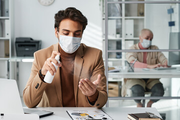 Young middle eastern employee in facial mask sitting at office table and applying spray disinfectant on hands