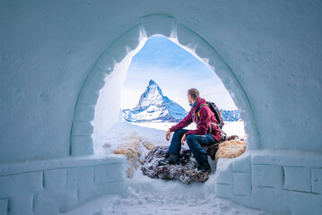 Male tourist sitting in entrance of igloo. Young man is looking at famous matterhorn peak against sky. He is in sportswear enjoying winter vacation in alps.