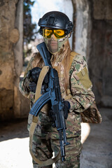 Fototapeta premium Portrait of a woman in a helmet and goggles with a machine gun in her hands. A female soldier in a camouflage uniform holds a weapon.