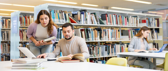 Positive young adults communicating while preparing to exam in library - obrazy, fototapety, plakaty