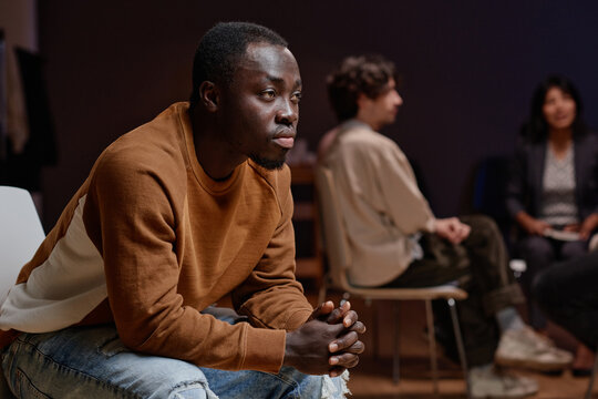 Horizontal medium shot of young Black man participating in group psychotherapy sitting on chair listening to someone