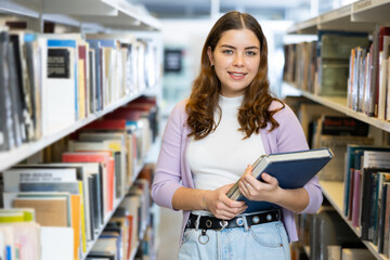 Portrait of young brown-haired woman holding stack of books, standing in library and looking at...