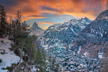 Snow covered houses against matterhorn mountain range during sunset. Scenic view of townscape by forest against cloudy sky. Idyllic view in alpine region during winter.