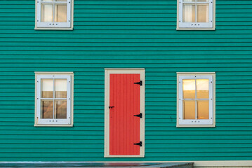 The exterior of a vibrant orange color single traditional wooden shutter door with cream color trim on a teal green clapboard wall of a historic house with four single hung windows with lace curtains.